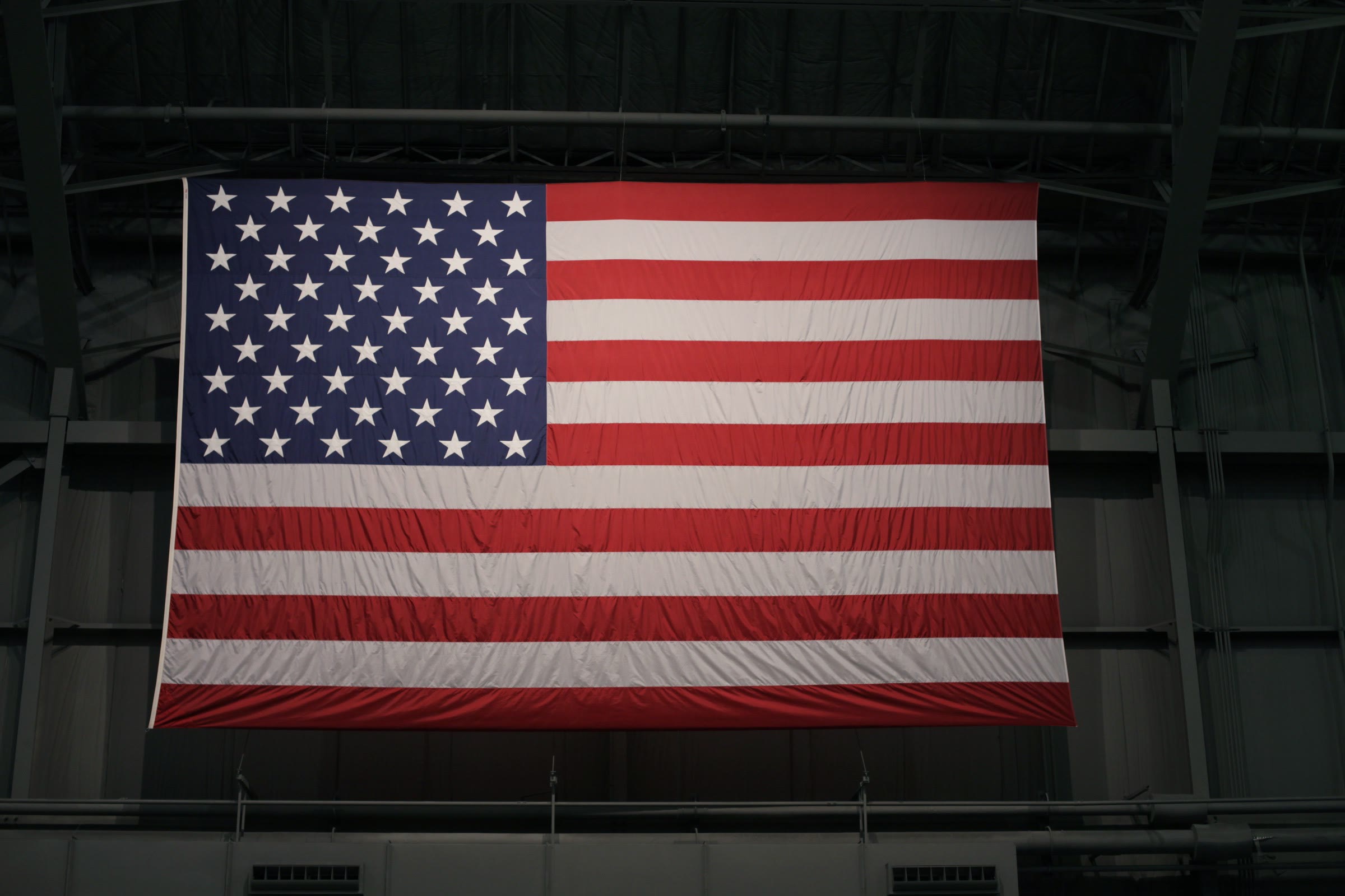 A large American flag hanging from the steel roof structure inside a manufacturing building