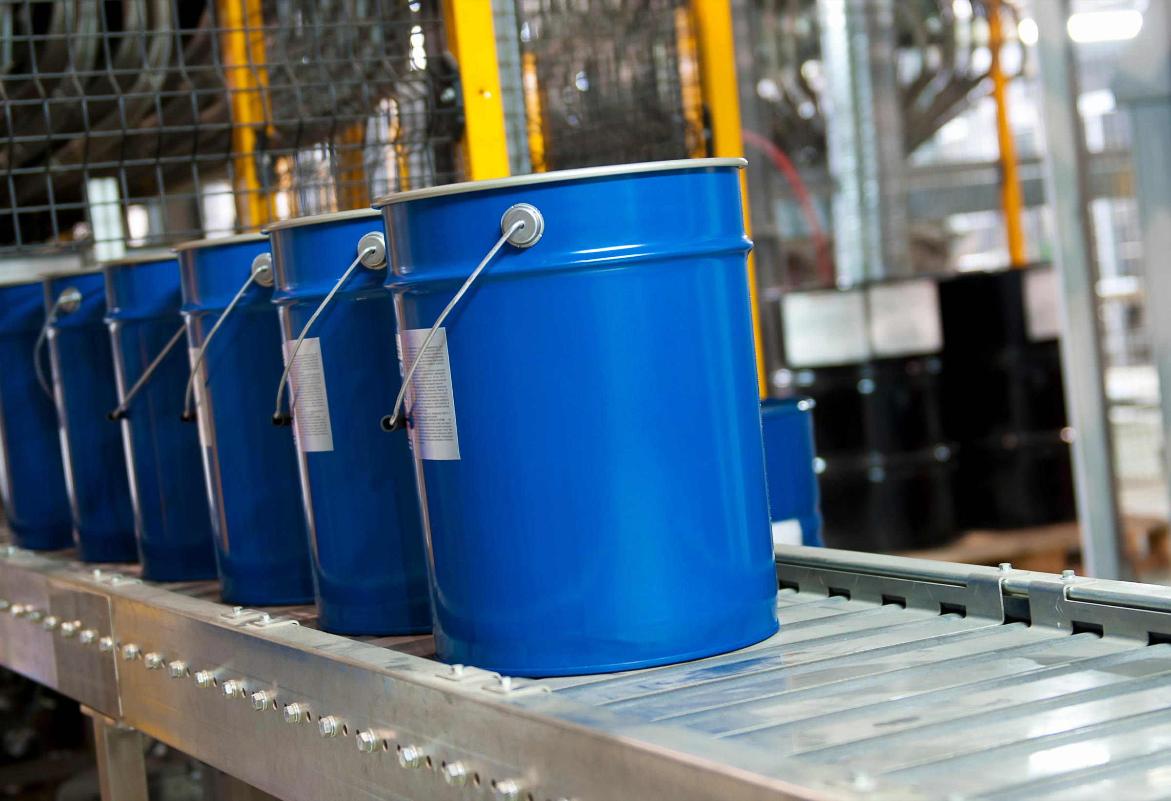 Blue steel pails with wire handles and paper hang tags moving along a roller conveyor in a coatings plant, black drums stacked behind