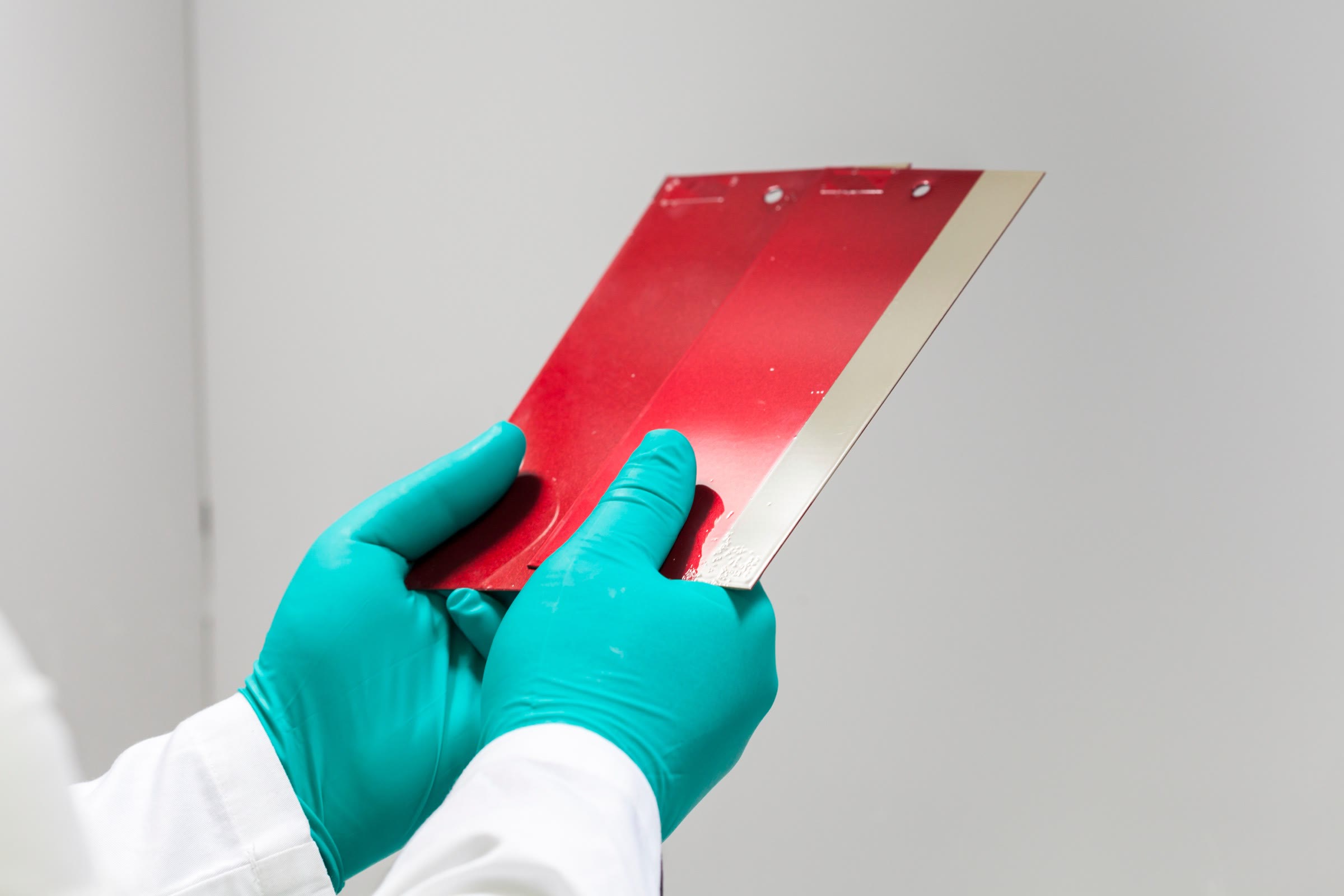Green-gloved hands in a white lab coat holding a red coated panel with a bare metal edge, against a plain light grey wall