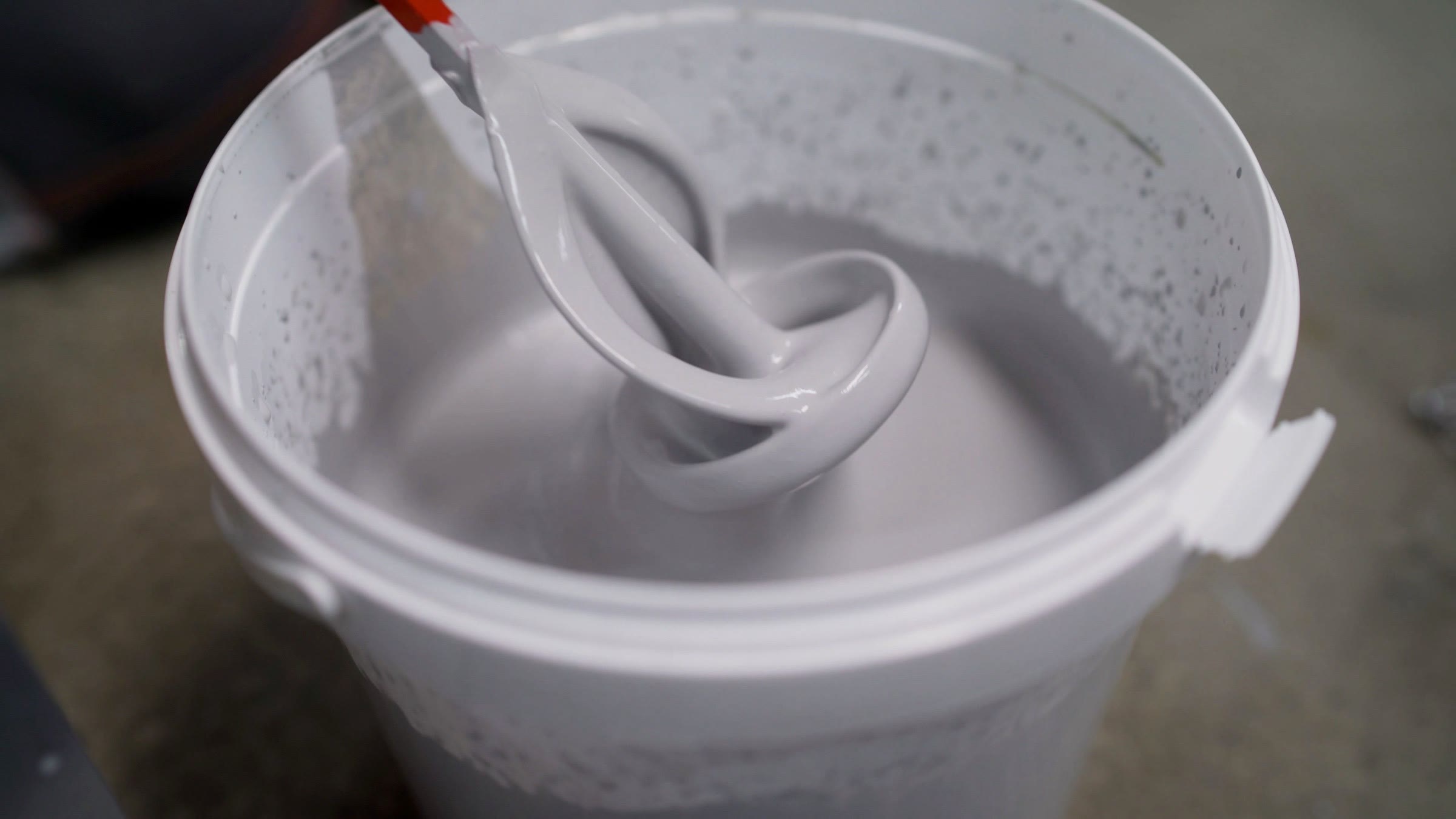 A paddle mixer on a drill folding thick grey coating in a plastic pail on a concrete floor