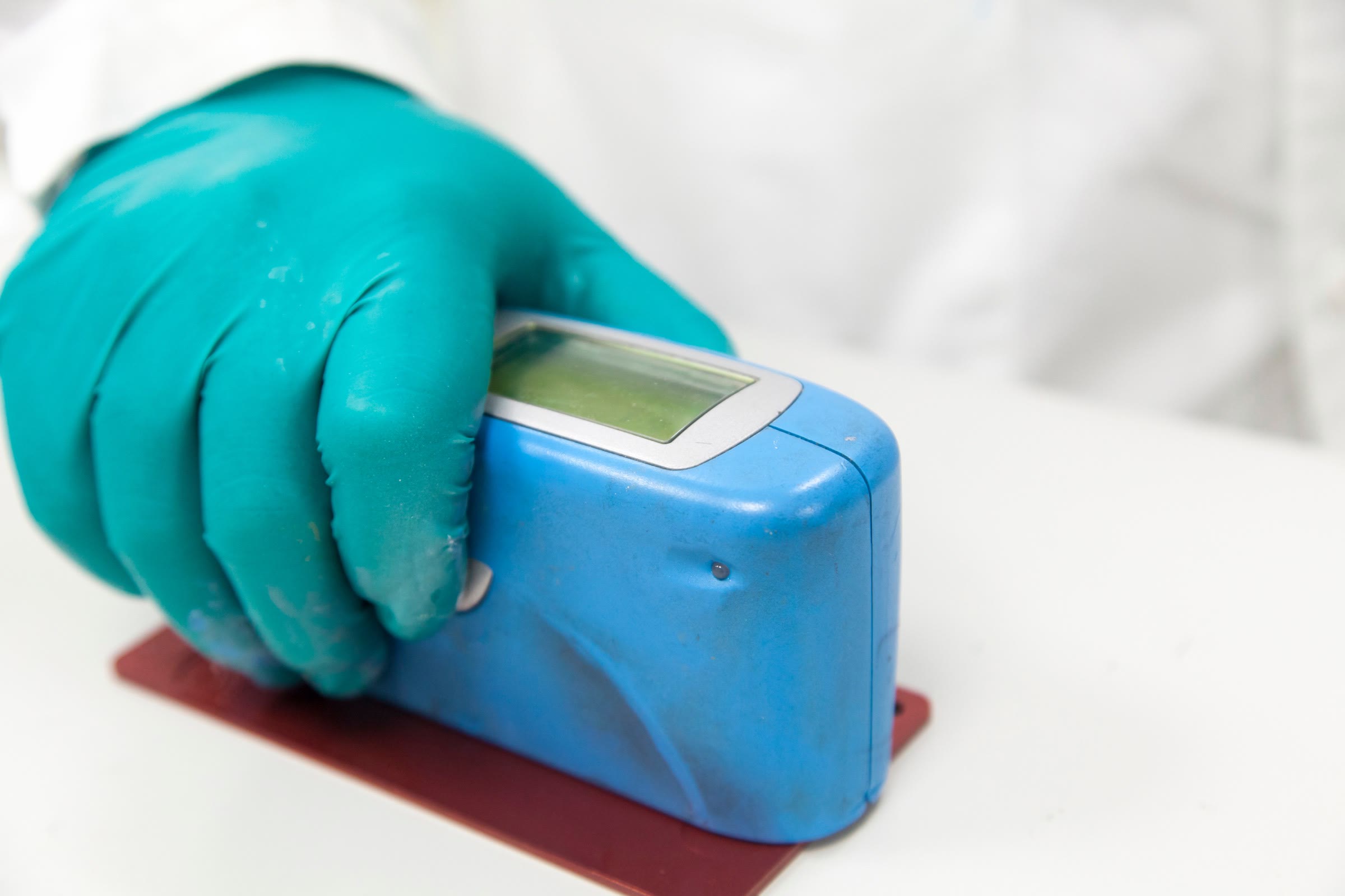 Gloved hand holding a handheld gloss meter on a dark red coated test panel