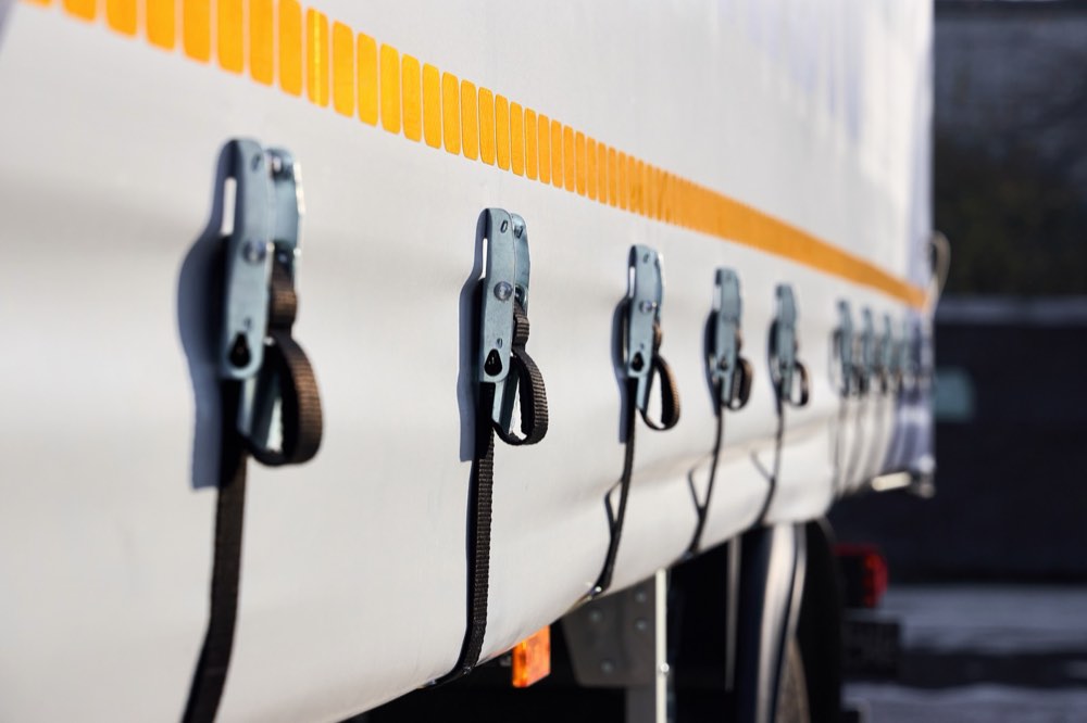Row of tensioning buckles and straps along the bottom edge of a white truck curtain